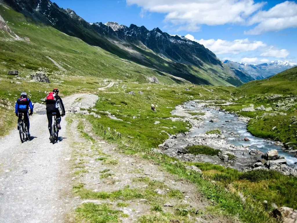 Dos personas en bicicleta de espaldas por una pista de montaña junto a un arroyo, con cumbres alpinas al fondo